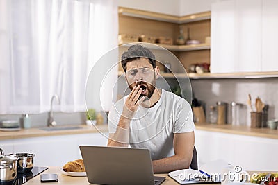 Sleepy tired bored handsome millennial caucasian guy with beard yawns, working at computer at table Stock Photo