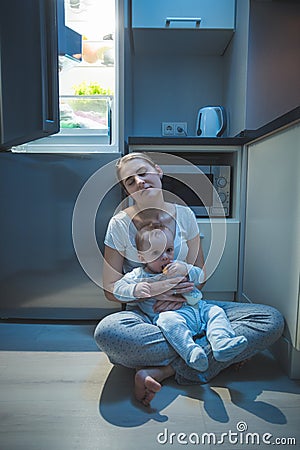 Sleepy tired woman sitting kitchen floor at night and feeding her baby boy with milk Stock Photo