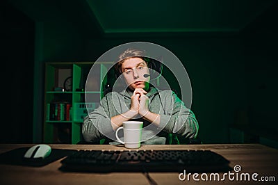 Sleepy young male gamer in headset sitting at night at the computer and looking at the camera with a tired face Stock Photo