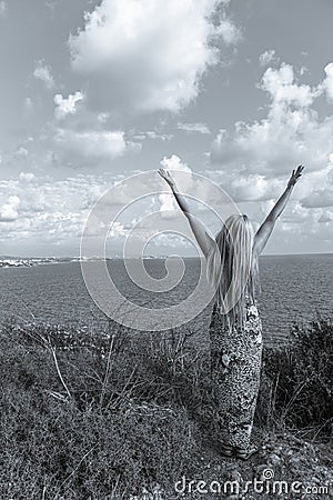 Slender young girl with long blonde hair on the mountain by the seashore with hands up to the sky. People connecting with nature Stock Photo