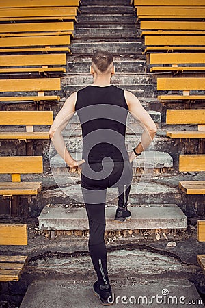 A slender young man in black sportswear stands in front of the stairs stadium stands. ready for action, step to victory, challenge Stock Photo