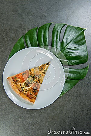 Slice of veggie pizza with monstera leaf under the serving plate. Piece of vegetable pizza with tomatoes, champignon mushrooms Stock Photo