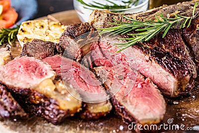 Sliced rare grilled or barbecued tomahawk beef steak on a griddle with fresh rosemary and tomato in a close up view. Stock Photo