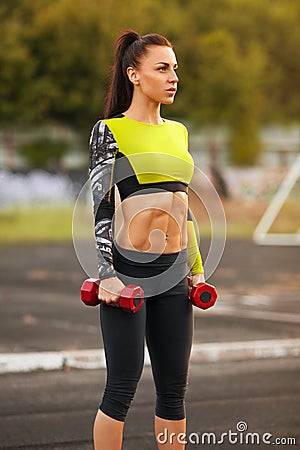 Slim athletic woman with dumbbells in the stadium. Sporty girl with flat belly workout, outdoors Stock Photo