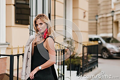 Slim blonde woman in trendy wristwatch standing near iron fence with gently smile posing in morning. Outdoor portrait of Stock Photo