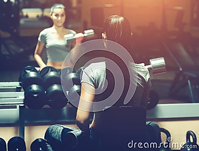 Slim, bodybuilder girl, lifts heavy dumbbell standing in front of the mirror while training in the gym. Stock Photo