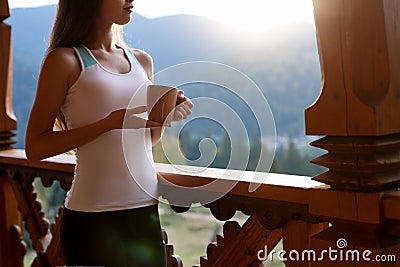 Slim caucasian woman holds cup of tea in her hands at mountain resort. Sports girl with hot coffee mug at wooden balcony Stock Photo