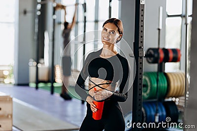 Slim dark-haired girl dressed in black sportswear stands with water in her hand near the sport equipment in the gym Stock Photo