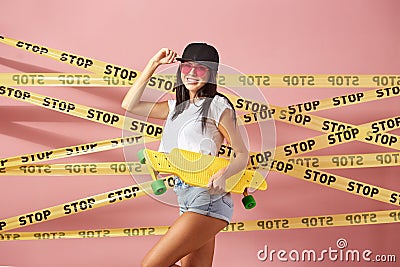 Slim dark-haired girl in pink sunglasses and cap dressed in jeans shorts and white top stands with yellow skateboard on Stock Photo