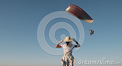 Slim girl stands and looks at the paraglider. Woman stands on a hill holding a hat in her hands and looks at a Stock Photo
