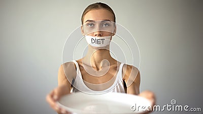 Slim girl with taped mouth showing empty plate, severe diet and self-destruction Stock Photo