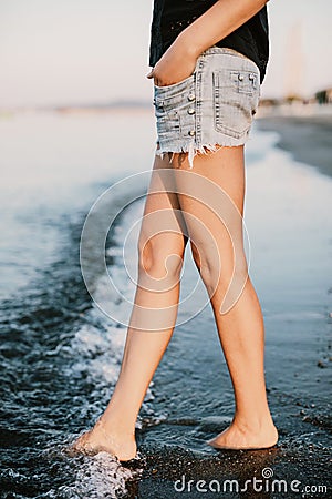Slim legs of a young and beautiful girl in shorts sit on the sand in the waves Stock Photo