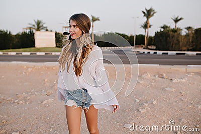 Slim long-haired girl in vintage white blouse walking on the sand with exotic palm trees on background. Charming young Stock Photo