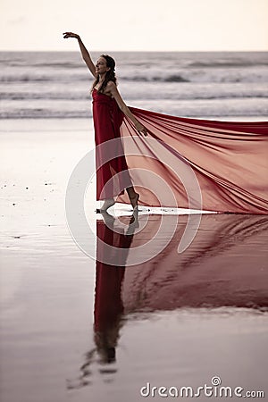 Slim woman dancing on the beach during sunset. Water reflection. Pretty Caucasian woman wearing long red dress. Perfect body shape Stock Photo