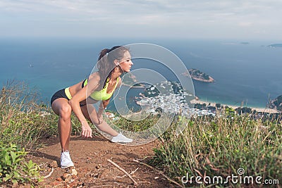 Slim young woman doing side lunge stretching exercise on top of the mountain with sea and sky in background Stock Photo