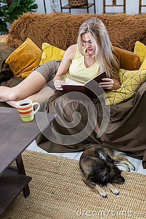 In the living room, a young woman is lying on the sofa reading a book. A dog is lying on the carpet. A multi-breed dog. Stock Photo