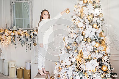 Small adorable female child in white sweater and trousers holds toy for decoration, decorates New Year tree. Cheerful little kid n Stock Photo