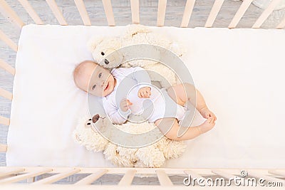 A small baby girl 6 months old in a white bodysuit is lying on her back in a child`s bed with soft toys bears Stock Photo