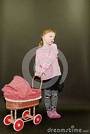 Small blond girl and dolls pram on black background. Little girl stands on black background. Stock Photo