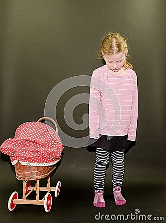 Small blond girl and dolls pram on black background. Little girl stands on black background. Stock Photo