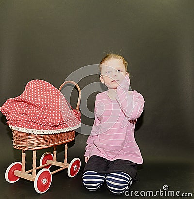 Small blond girl and dolls pram on black background. Little girl sits on black background. Stock Photo