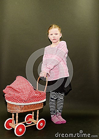 Small blond girl and dolls pram on black background. Little girl stands on black background. Stock Photo