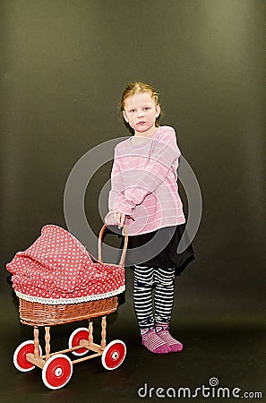 Small blond girl and dolls pram on black background. Little girl stands on black background. Stock Photo