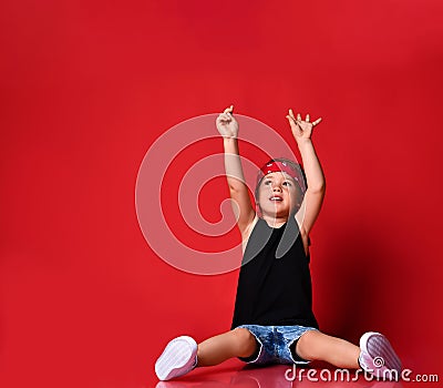 Small boy in stylish casual clothing, hair bandana and white sneakers sitting on floor and feeling excited with hands raised up Stock Photo