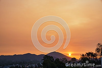 Small bright sun in light orange sunset sky shining behind mountains above houses and green trees Stock Photo