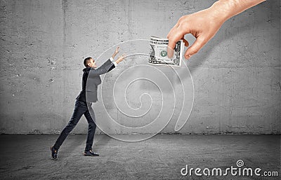 Small businessman on concrete background reaching out for a giant hand holding money. Stock Photo