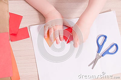 A small child glues paper, children`s safe scissors and colored paper on the table Stock Photo