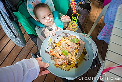 A small child at 8 months wants to eat adult food and pulls a plate of shrimps and vegetables to him. Baby Sitting in a stroller Stock Photo