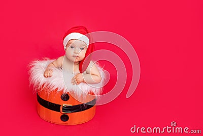 A small child of six months in a Santa hat sits in a basket on a red isolated photophone, a place for text, the concept of the new Stock Photo