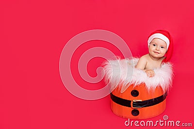 A small child of six months in a Santa hat sits in a basket on a red isolated photophone, a place for text, the concept of the new Stock Photo