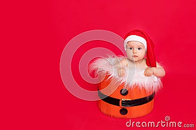 A small child of six months in a Santa hat sits in a basket on a red isolated photophone, a place for text, the concept of the new Stock Photo