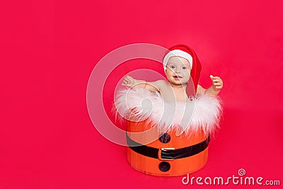 A small child of six months in a Santa hat sits in a basket on a red isolated photophone, a place for text, the concept of the new Stock Photo