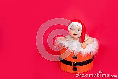 A small child of six months in a Santa hat sits in a basket on a red isolated photophone, a place for text, the concept of the new Stock Photo