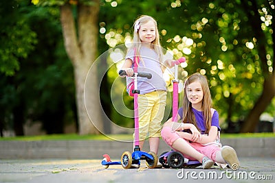 Small children learning to ride scooters in a city park on sunny summer evening. Cute little girls riding rollers. Stock Photo