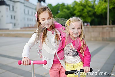 Small children learning to ride scooters in a city park on sunny summer evening. Cute little girls riding rollers. Stock Photo