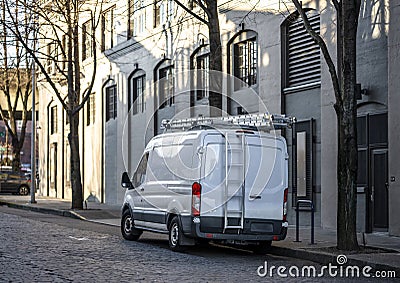 Small compact cargo mini van with ladder on the roof standing on the city street with multilevel buildings Stock Photo