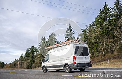 Small compact commercial cargo mini van with ladders on the roof running on the road with trees on the hill Stock Photo