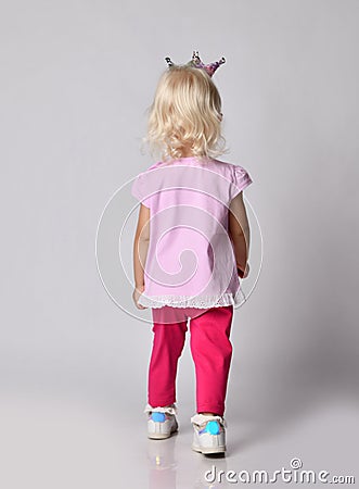 Small cute blonde baby girl kid in pink stylish casual clothing and crown stands back to camera, leaving out Stock Photo