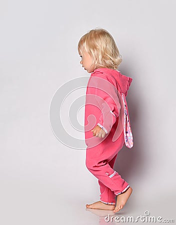 Small cute blonde baby girl in pink warm comfortable jumpsuit walking over grey wall background Stock Photo