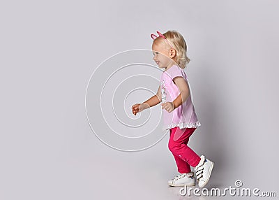 Small cute blonde smiling baby girl in pink stylish casual clothing and kitty ears running towards free copy space Stock Photo