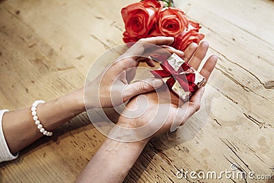 Small cute present box with bow in beautiful woman hands. Focus on bow. Red roses flowers behind on wooden table. St. Valentine`s Stock Photo