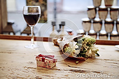 Small cute present box with bow at wooden table, flowers and glass of wine behind. Romantic meeting in cafe. St. Valentine`s day c Stock Photo