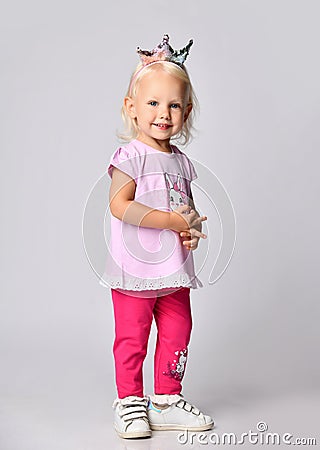 Small cute smiling blonde baby girl kid in pink stylish casual clothing and princess crown stands looking at camera Stock Photo