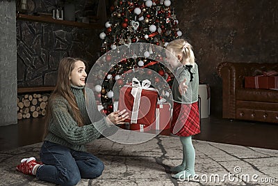 Small daughter gives big gift to her mom near Christmas tree. Portrait of mother and daughter on New Year Eve Stock Photo