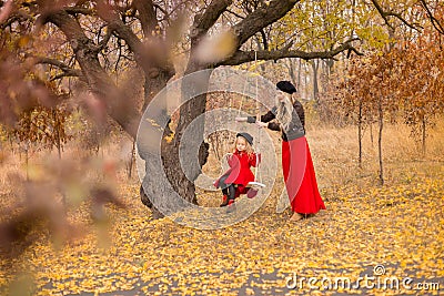 Beautiful mother in a long dress is swinging on a hinged swing little daughter in a red coat in an autumn garden near an old gnarl Stock Photo