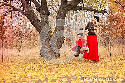 Beautiful mother in a long dress is swinging on a hinged swing little daughter in a red coat in an autumn garden near an old gnarl Stock Photo